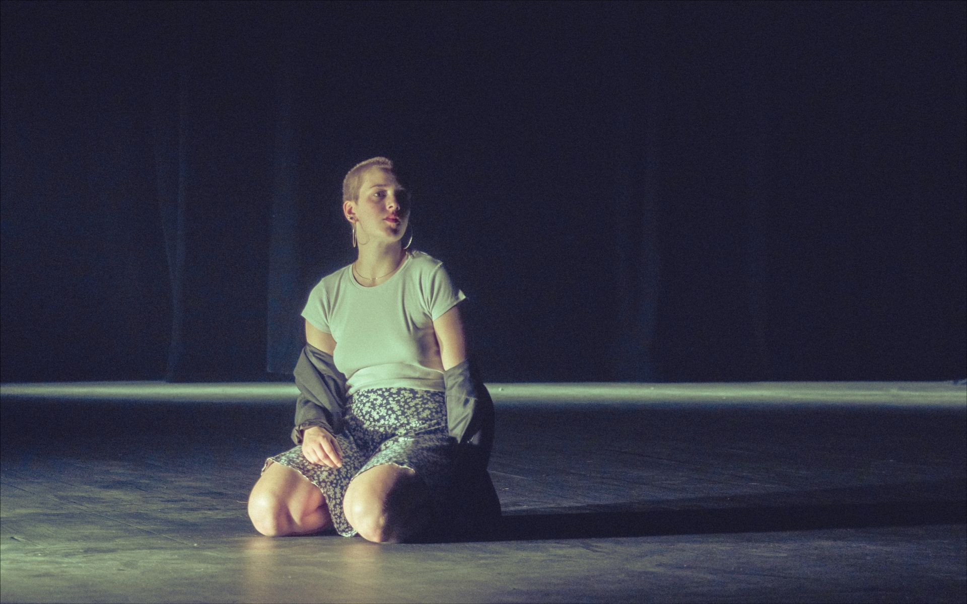 Woman kneeling on a stage, lit by a single beam. Wearing a t-shirt and skirt, jacket draped over shoulders.