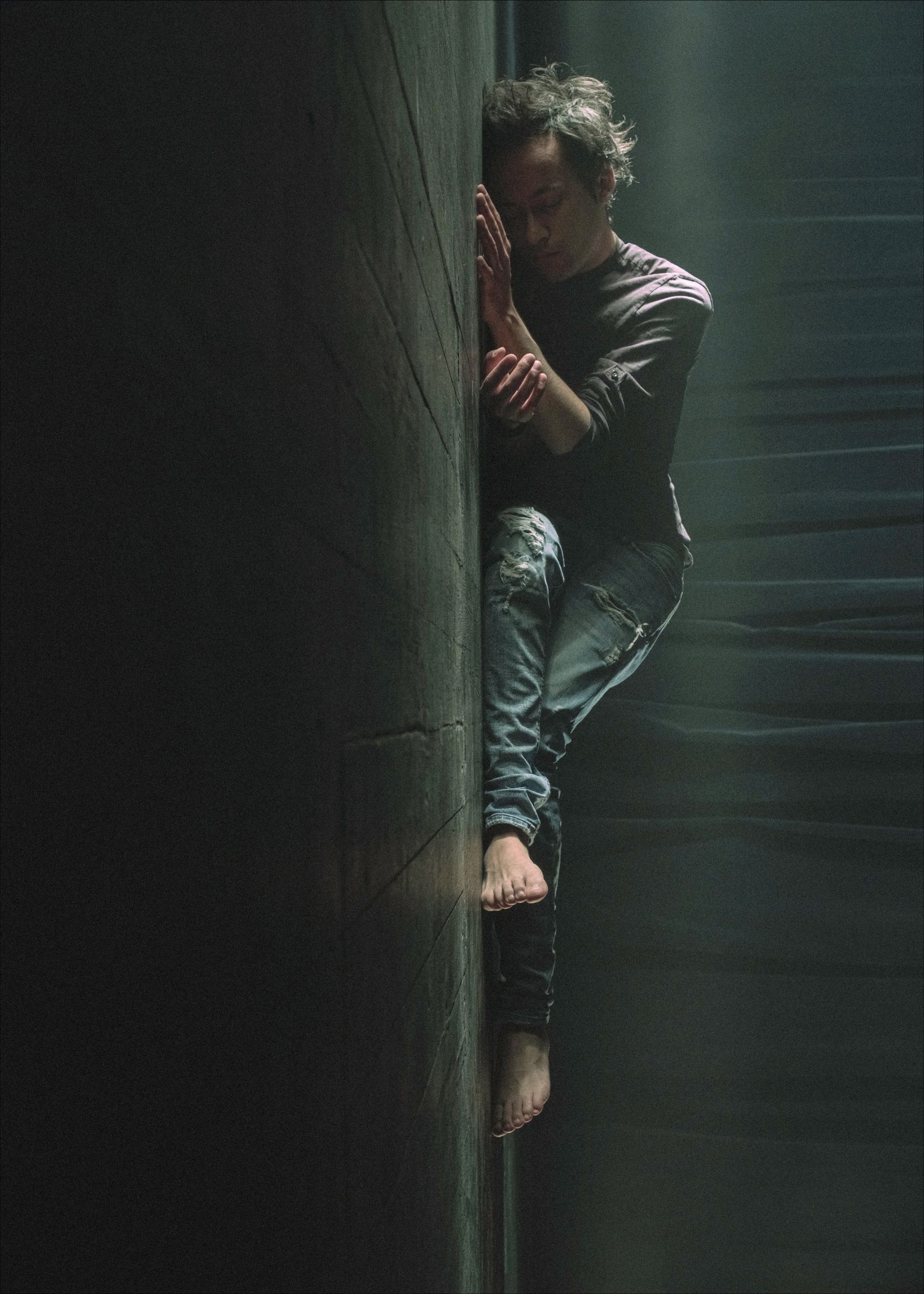 Man clinging to a dark wall, feet dangling. Barefoot, holding face, appears despondent, in shadowy space.