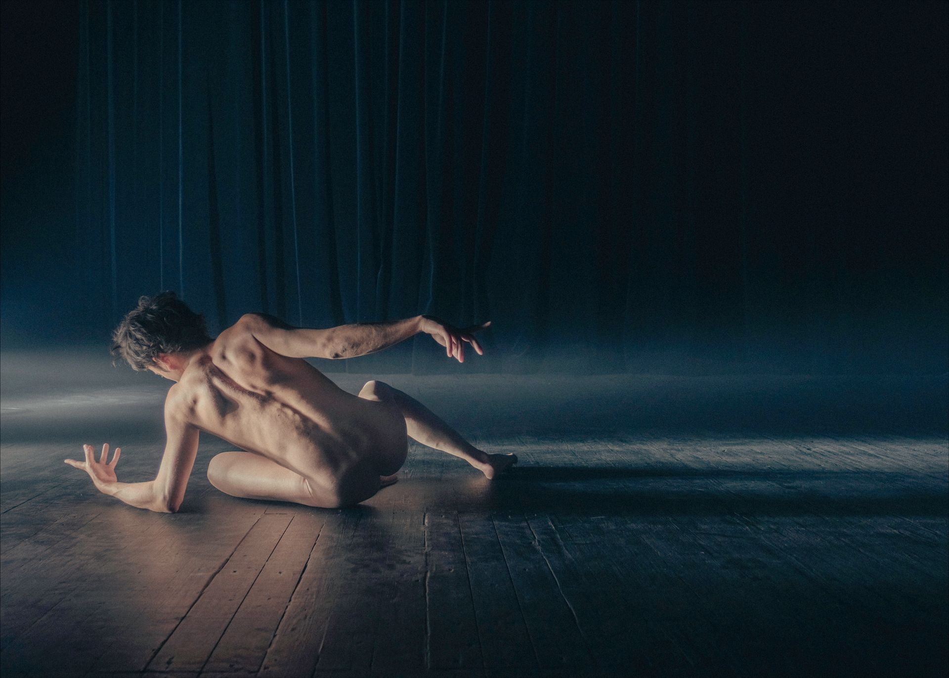 Naked dancer on wooden floor, backlit, reaching with arms, dramatic lighting, dark background.