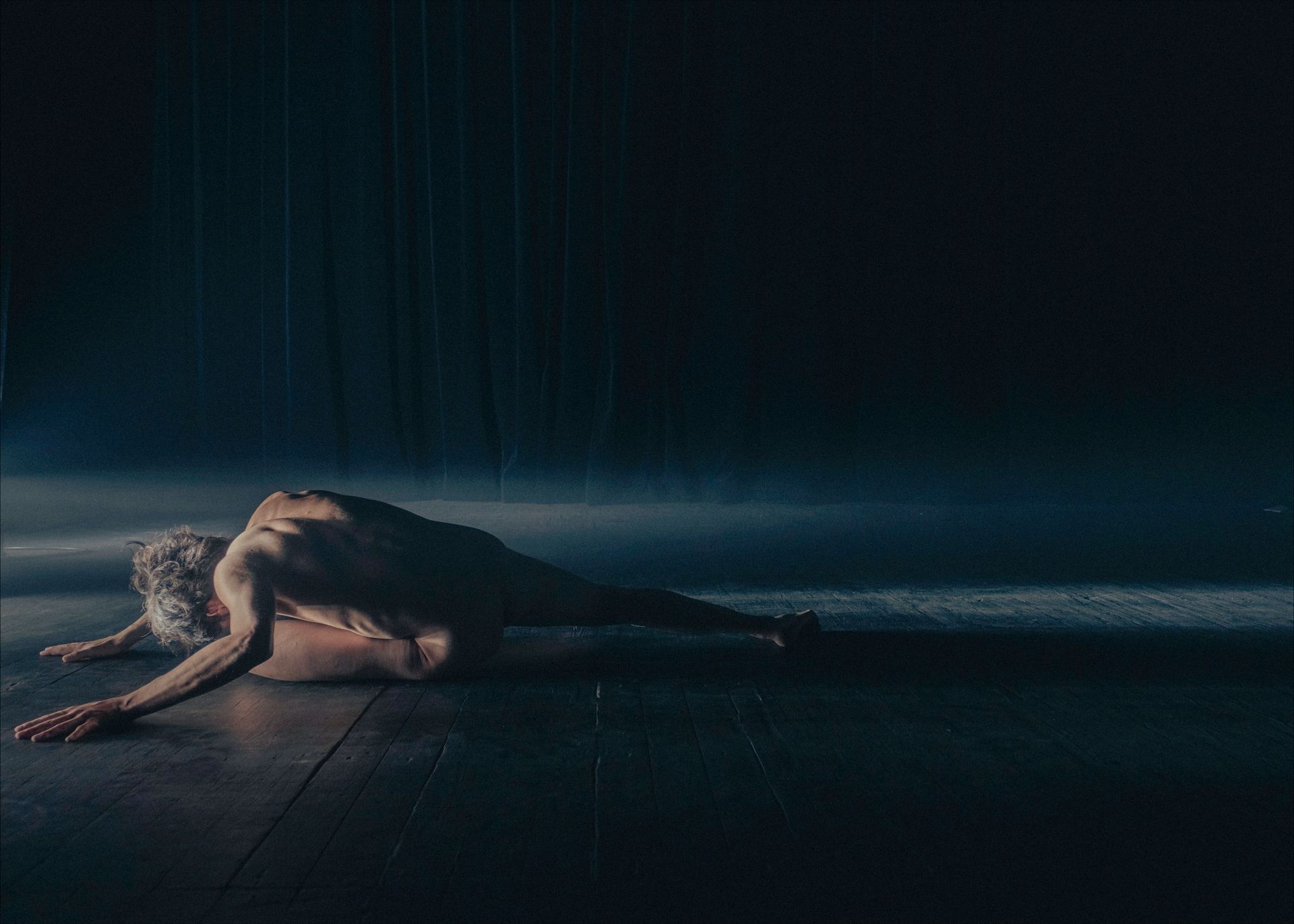 A figure lies on a dark floor with outstretched arm; lit by a single light, falling rain.