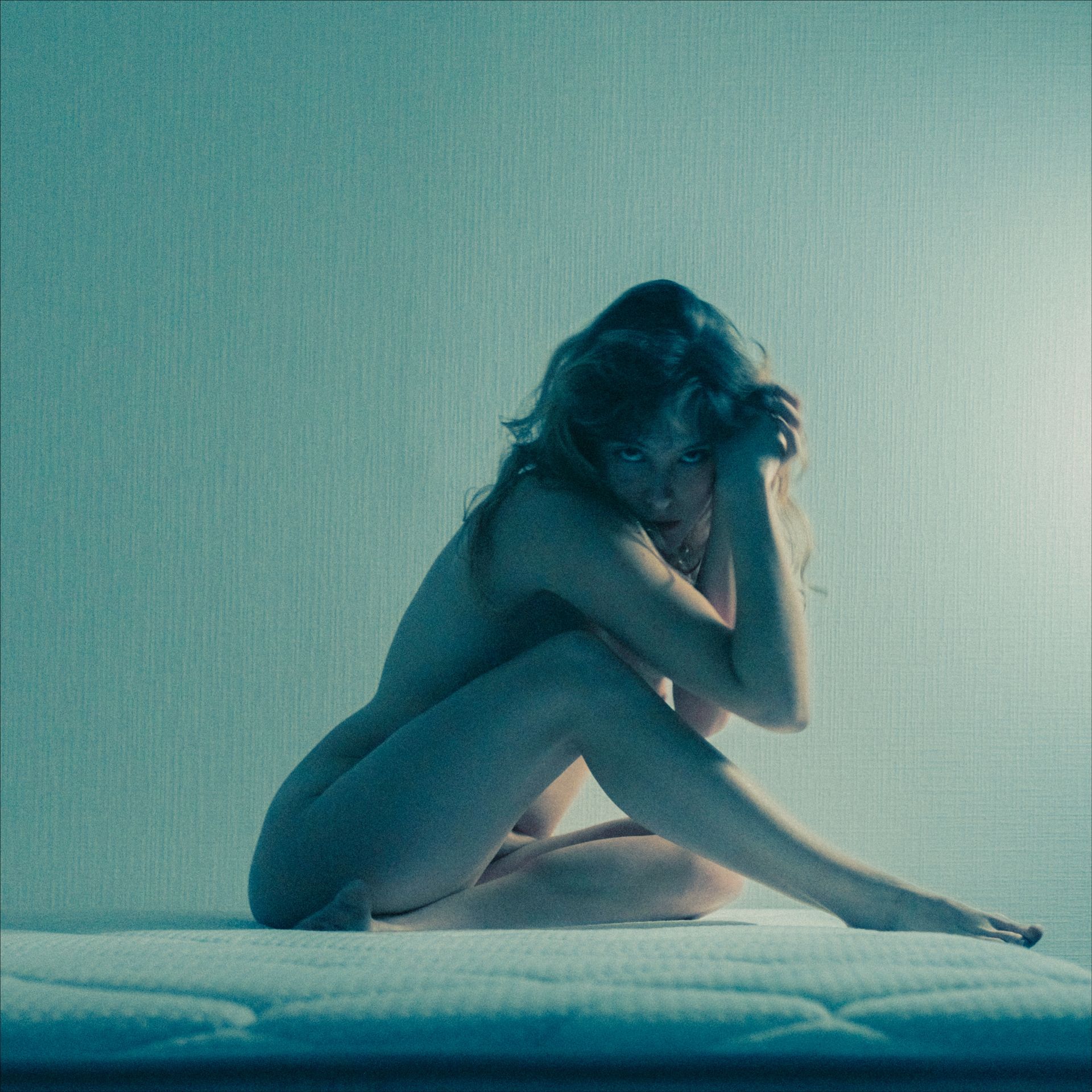 Naked person sitting on a mattress, head in hands, dimly lit, cyan-toned room.
