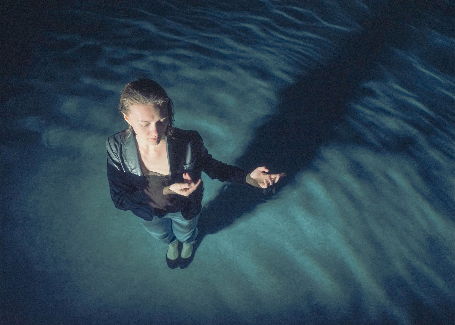 Woman standing in blue-lit water with arms outstretched, casting a long shadow.