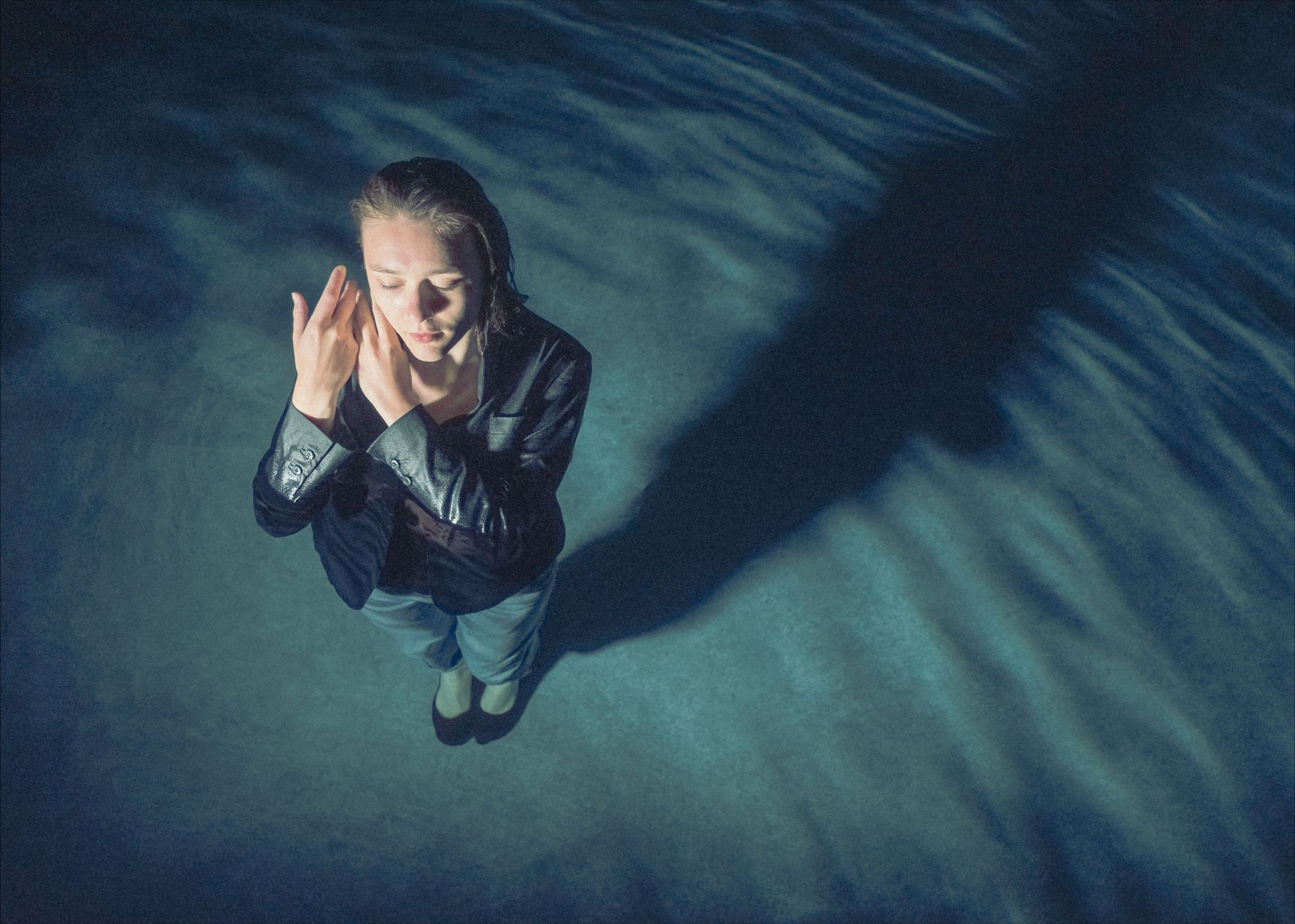 Woman standing in water, hands to face, looking down, shadowed in blue light.