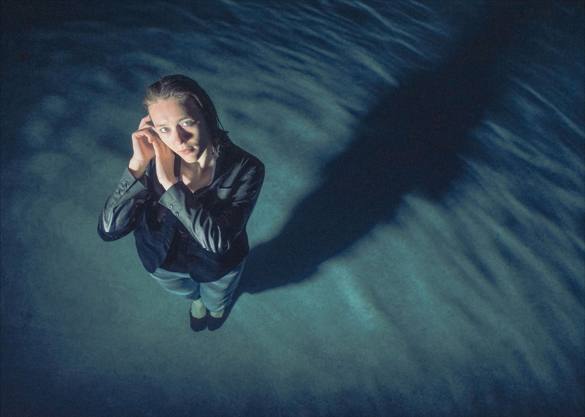 Woman in a wet jacket stands in dark water, hands near her face, looking up. Long shadow.