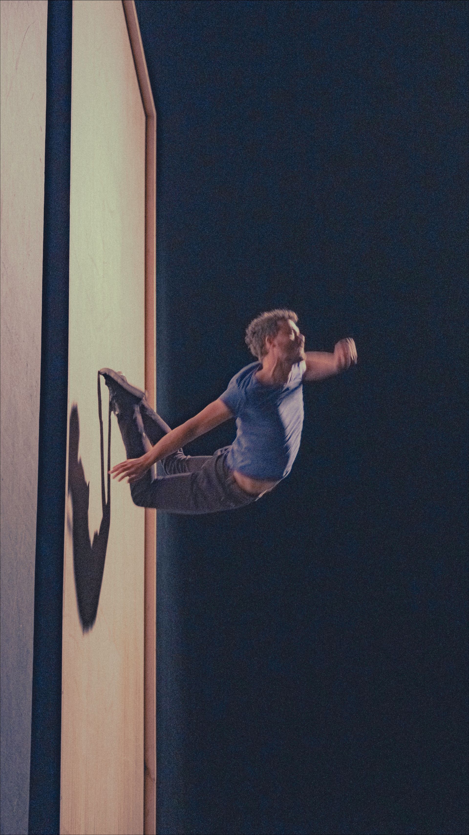 Man in blue shirt, in mid-air, appears to be exiting a wall. Dark setting with a shadow.