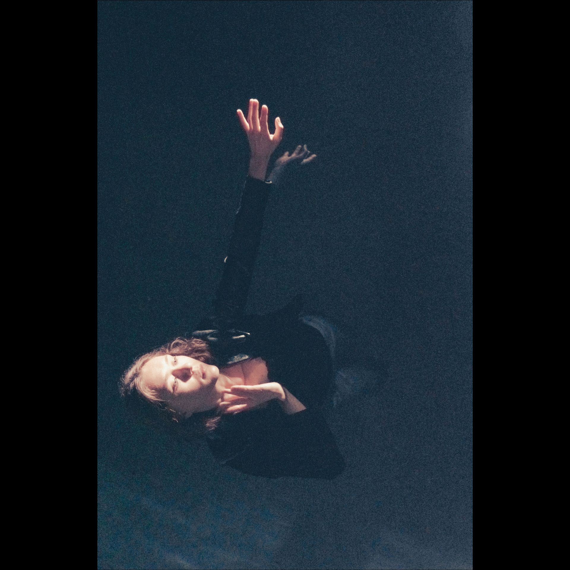 Person with arms raised, lit against a dark background, in a dance pose.