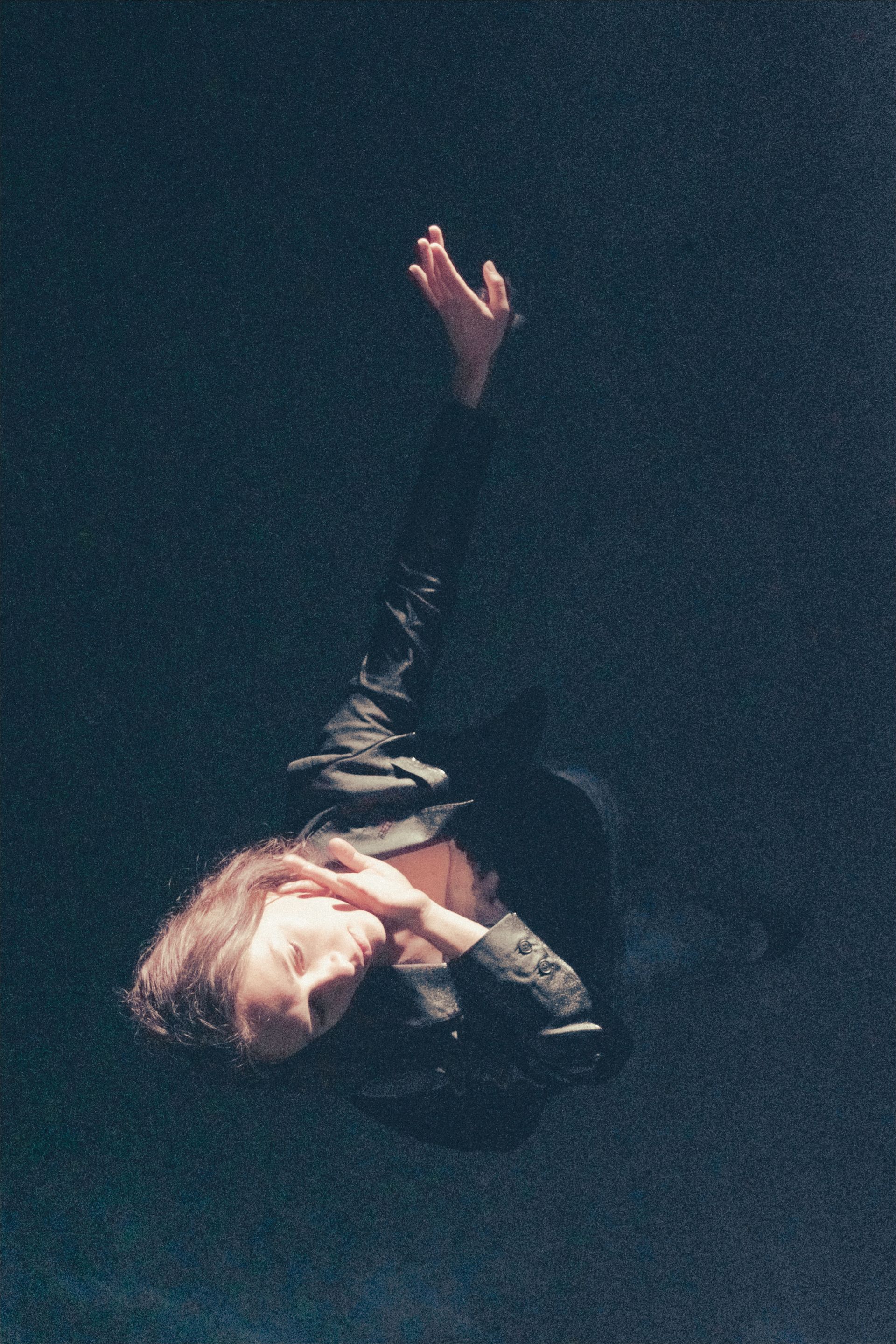 Person in dark clothing, arms outstretched upward, eyes closed. Dark, grainy background.