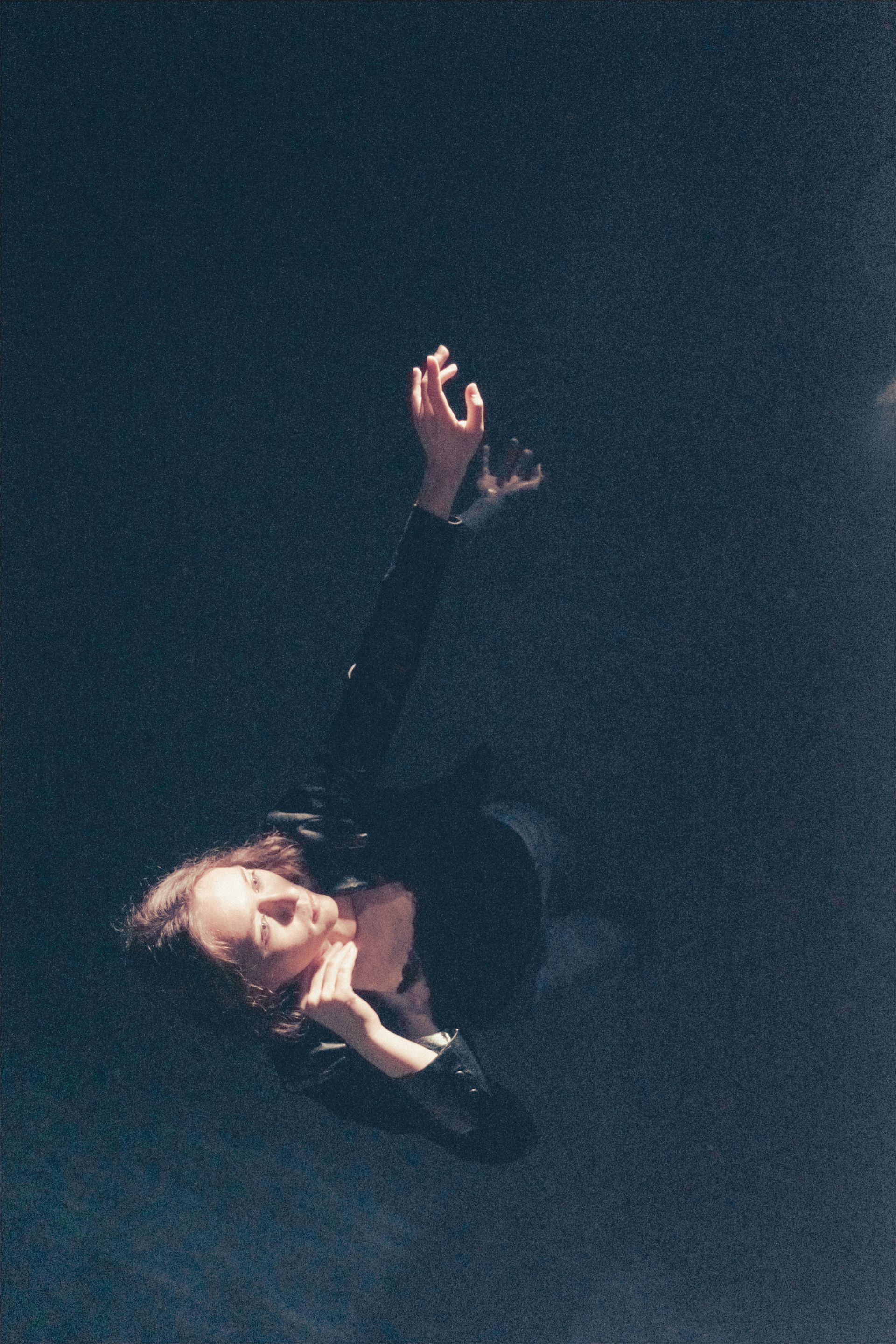 Person floating in a dark space, arms raised and head tilted back, soft lighting.