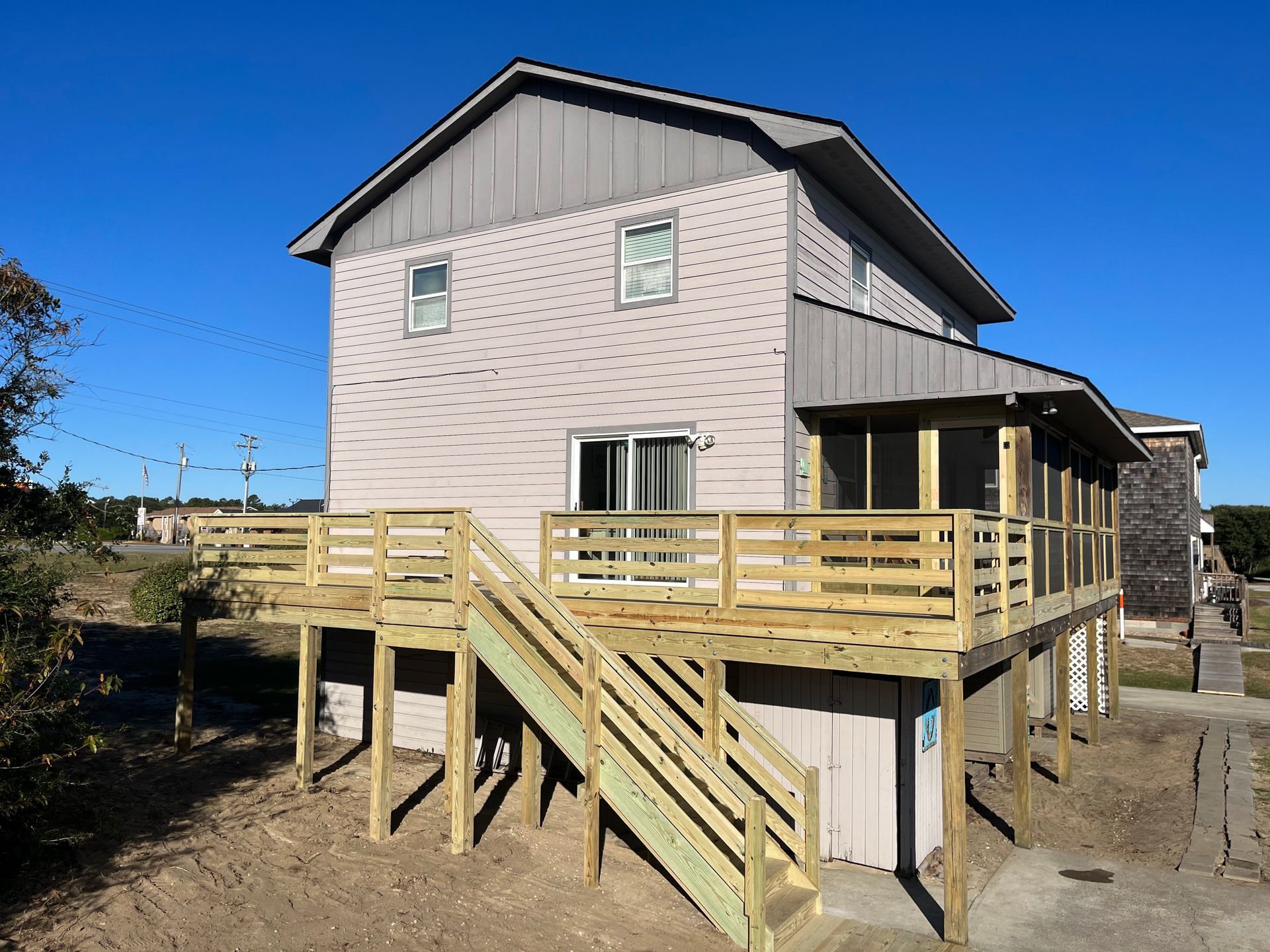 Two-story light gray house with wooden deck and stairs, blue sky.