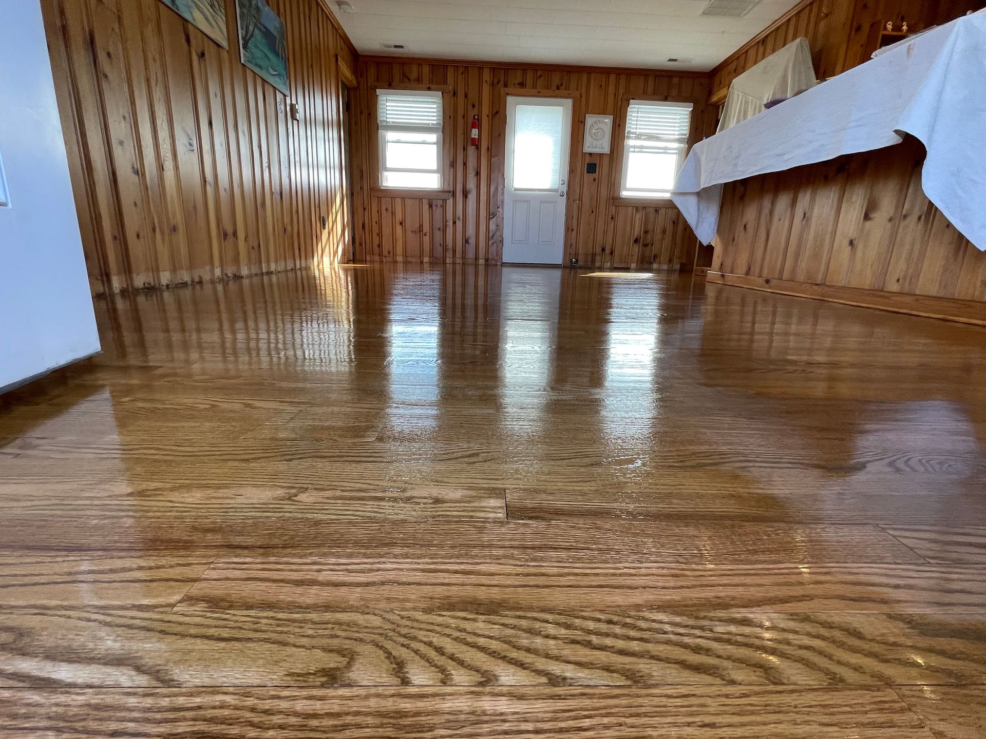 Shiny wooden floor reflecting natural light in a room with wood-paneled walls, windows, and a white tablecloth.