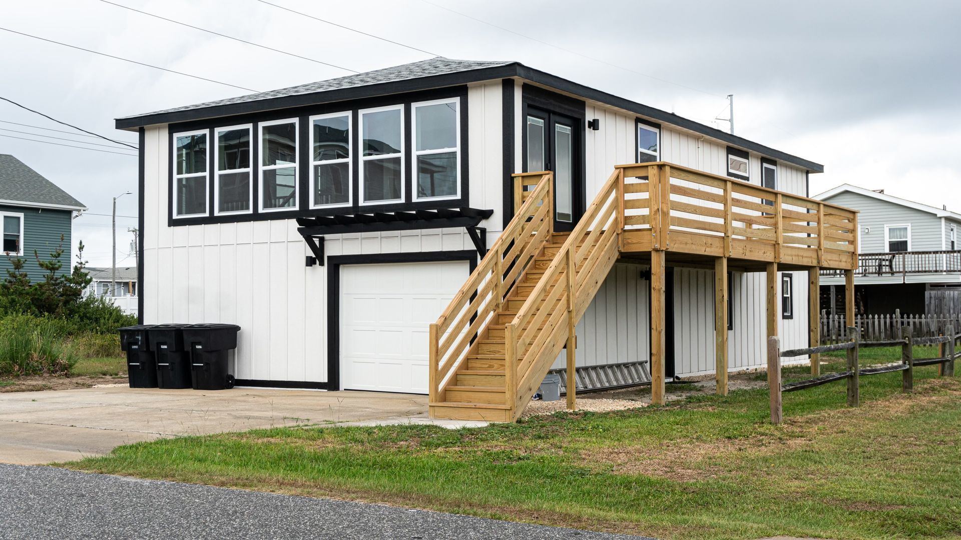 White two-story house with black trim, a garage, and wooden staircase. Three trash bins sit in the driveway.