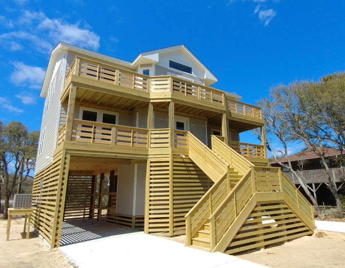 Beach house with wooden decks and stairs, gray and white siding, on a sunny day.