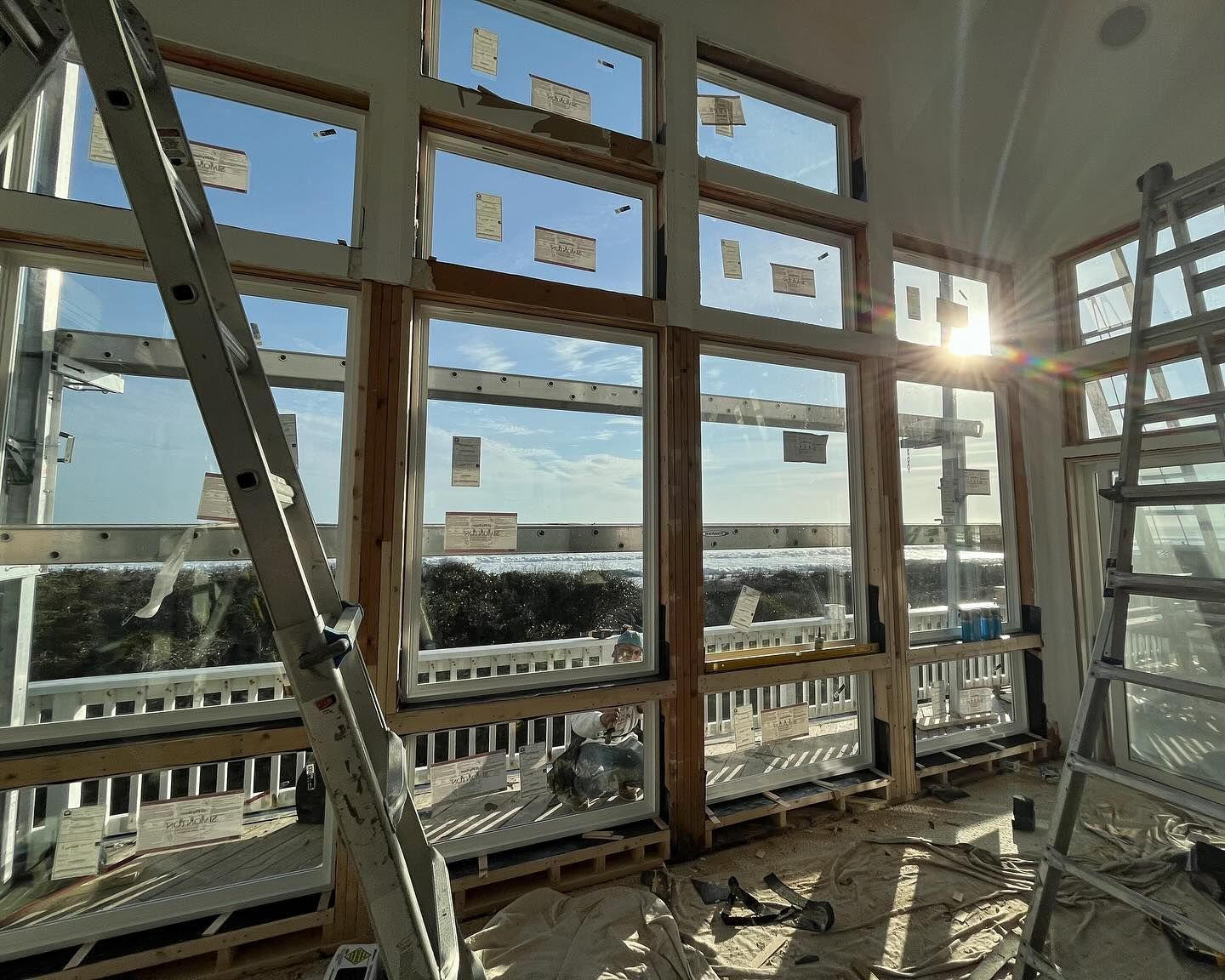 Interior view of a room with large windows under construction, sunlight streaming through. Ladder leaning against wall.