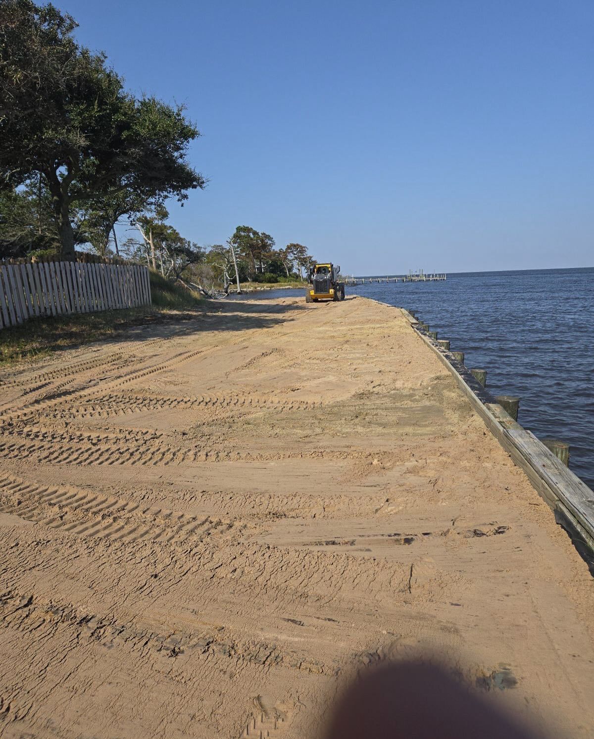 Sandy shoreline with tire tracks; small construction vehicle; water, blue sky.