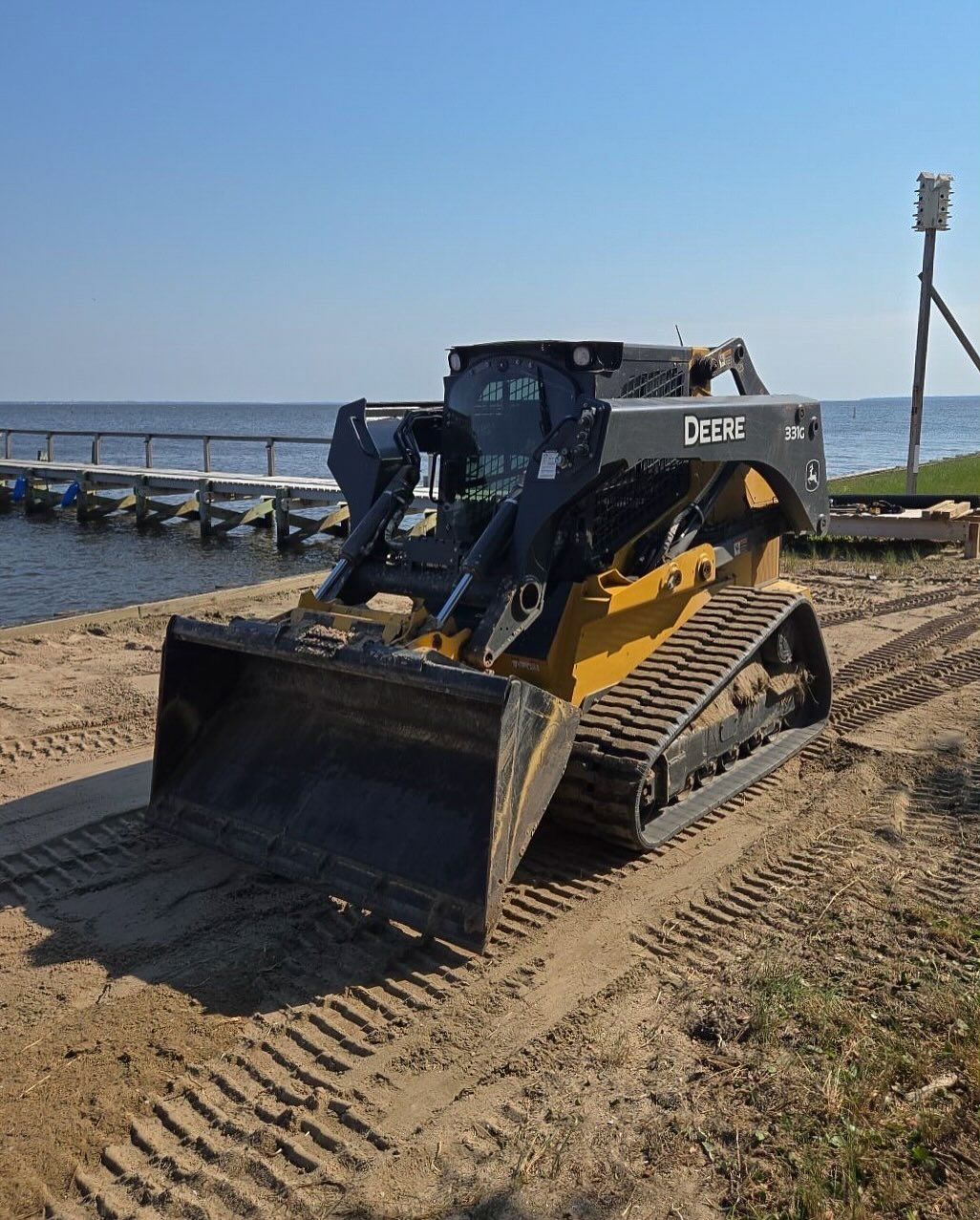 Yellow and black skid steer on sand by water, tracks visible.