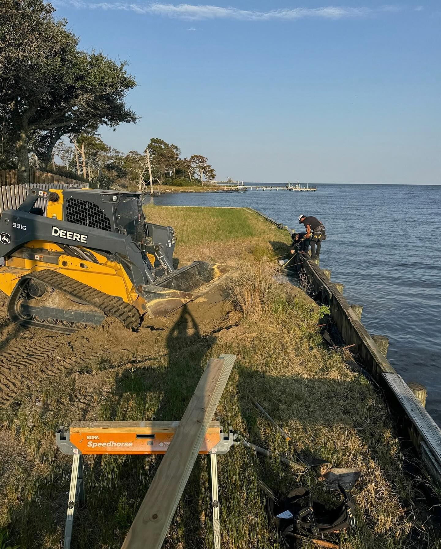 A person repairs a waterside retaining wall, with a yellow tractor and calm water in view.