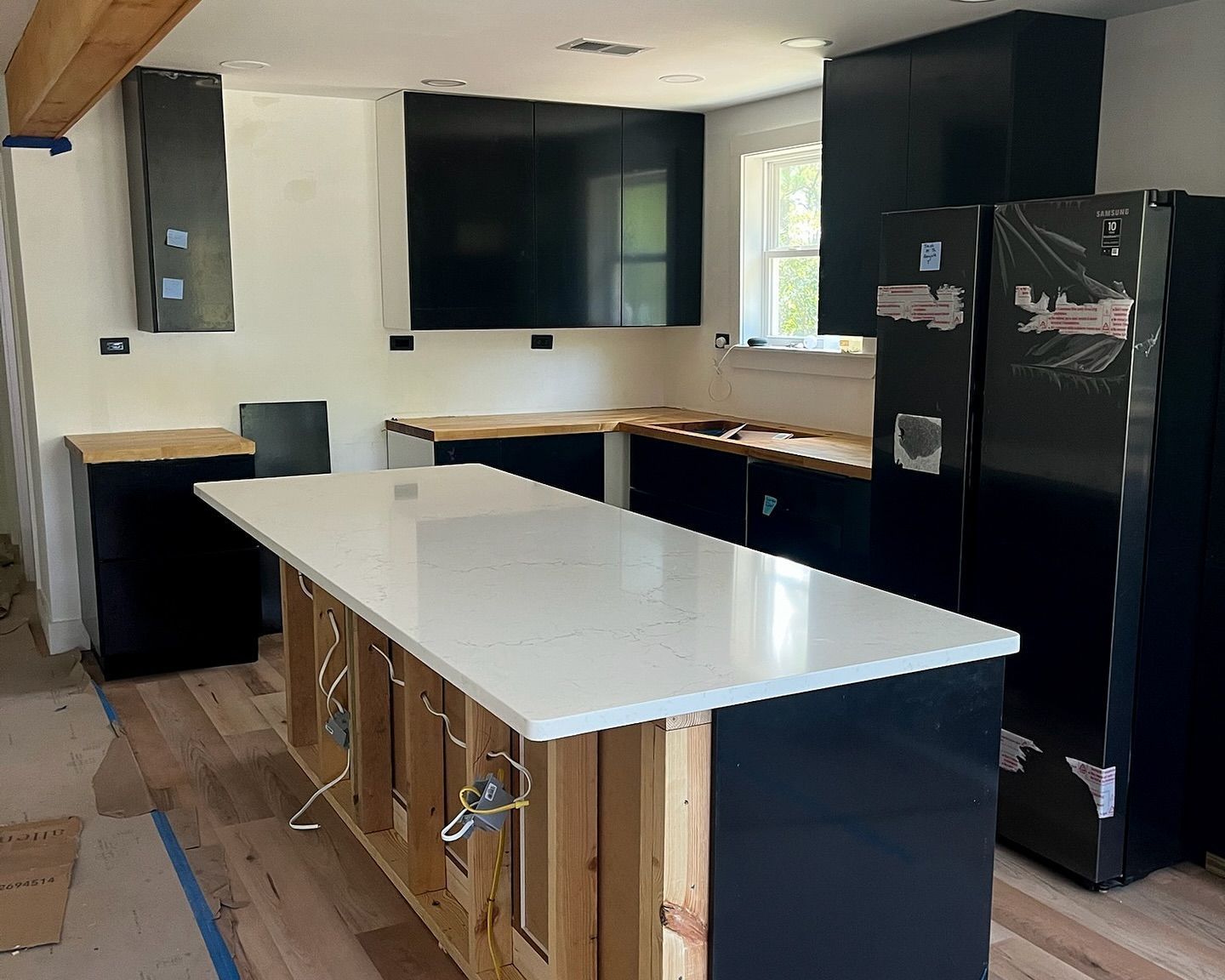 Kitchen under construction: black cabinets, white island countertop, light wood floors, a large TV.