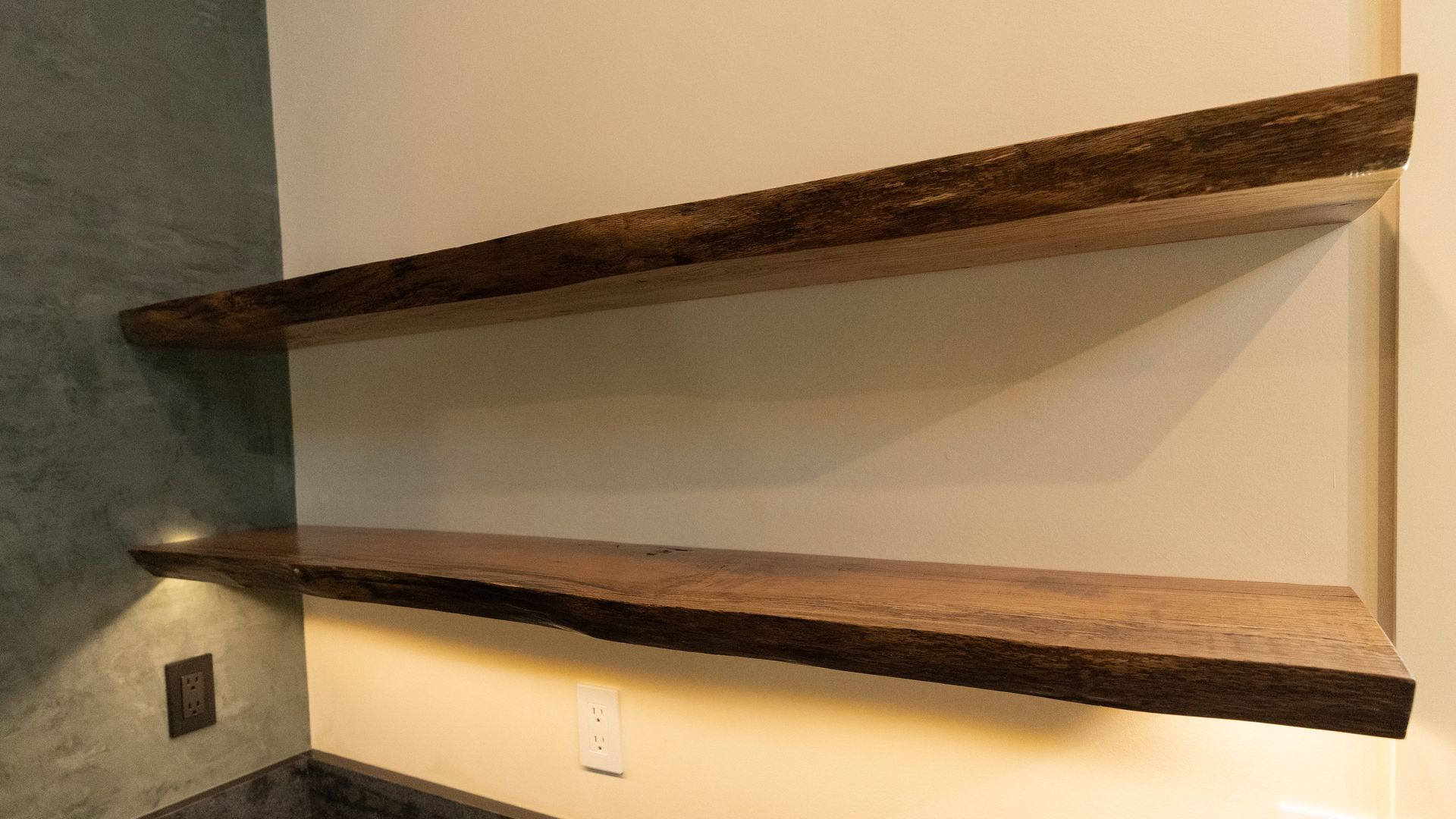 Two dark, wooden floating shelves mounted on a light beige wall with under-shelf lighting.