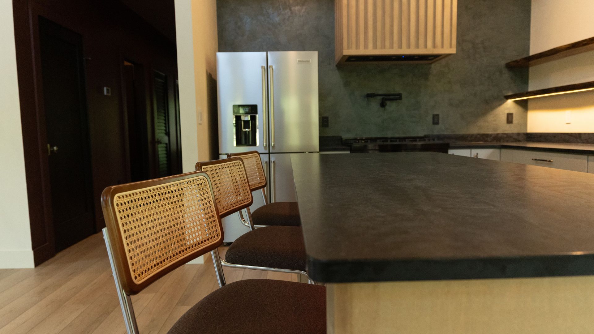 Modern kitchen with island and rattan chairs, stainless steel fridge.