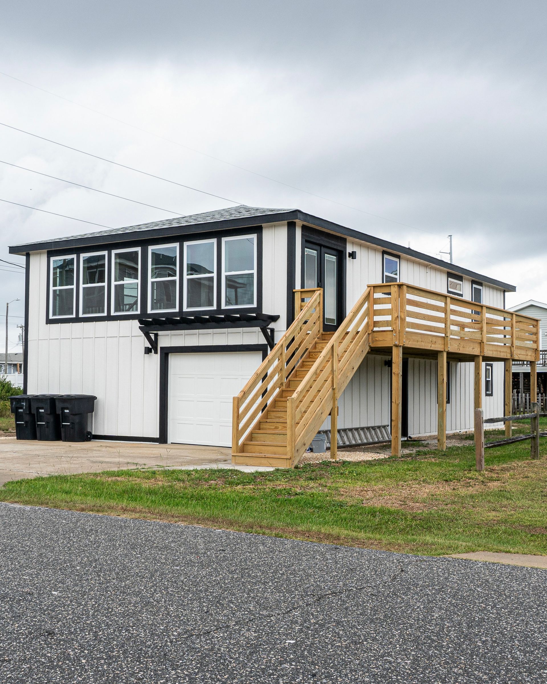 White house with black trim, wooden deck, stairs, and a garage on a gravel driveway under cloudy skies.