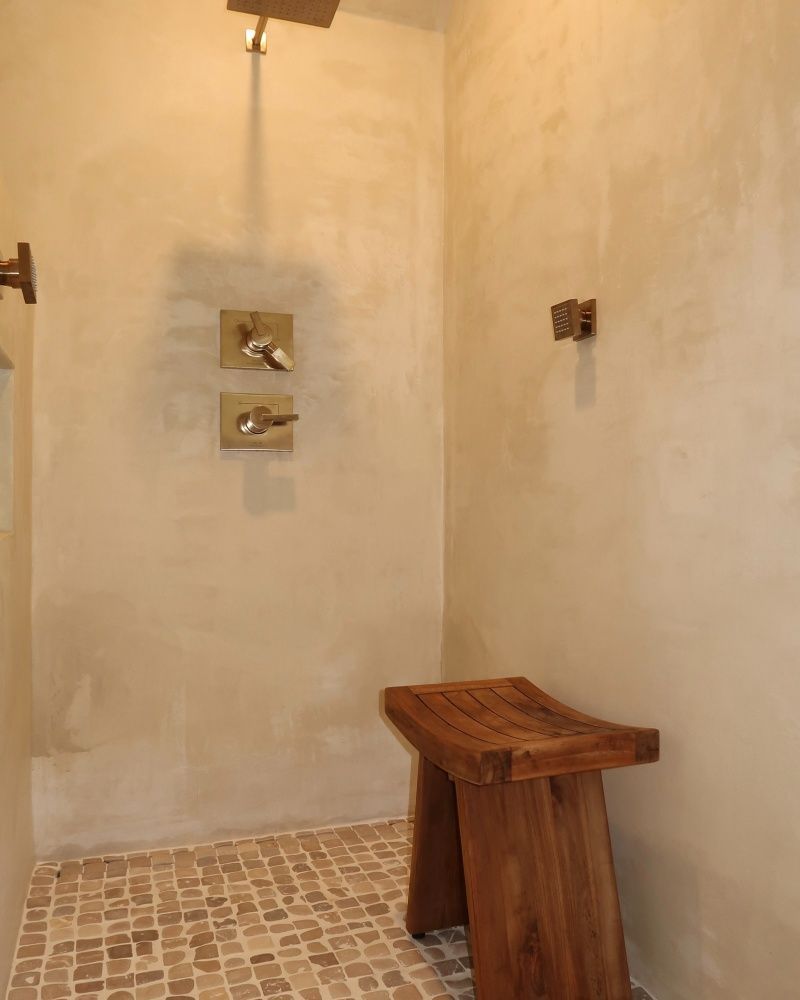 Shower stall with beige walls, a wooden stool, and small tiled floor. Brass fixtures and a shower head are mounted.