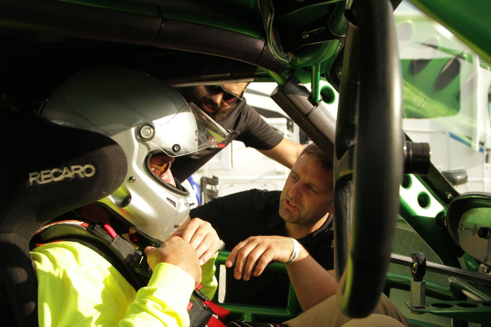A race car driver in helmet is checked by two men inside a green car.