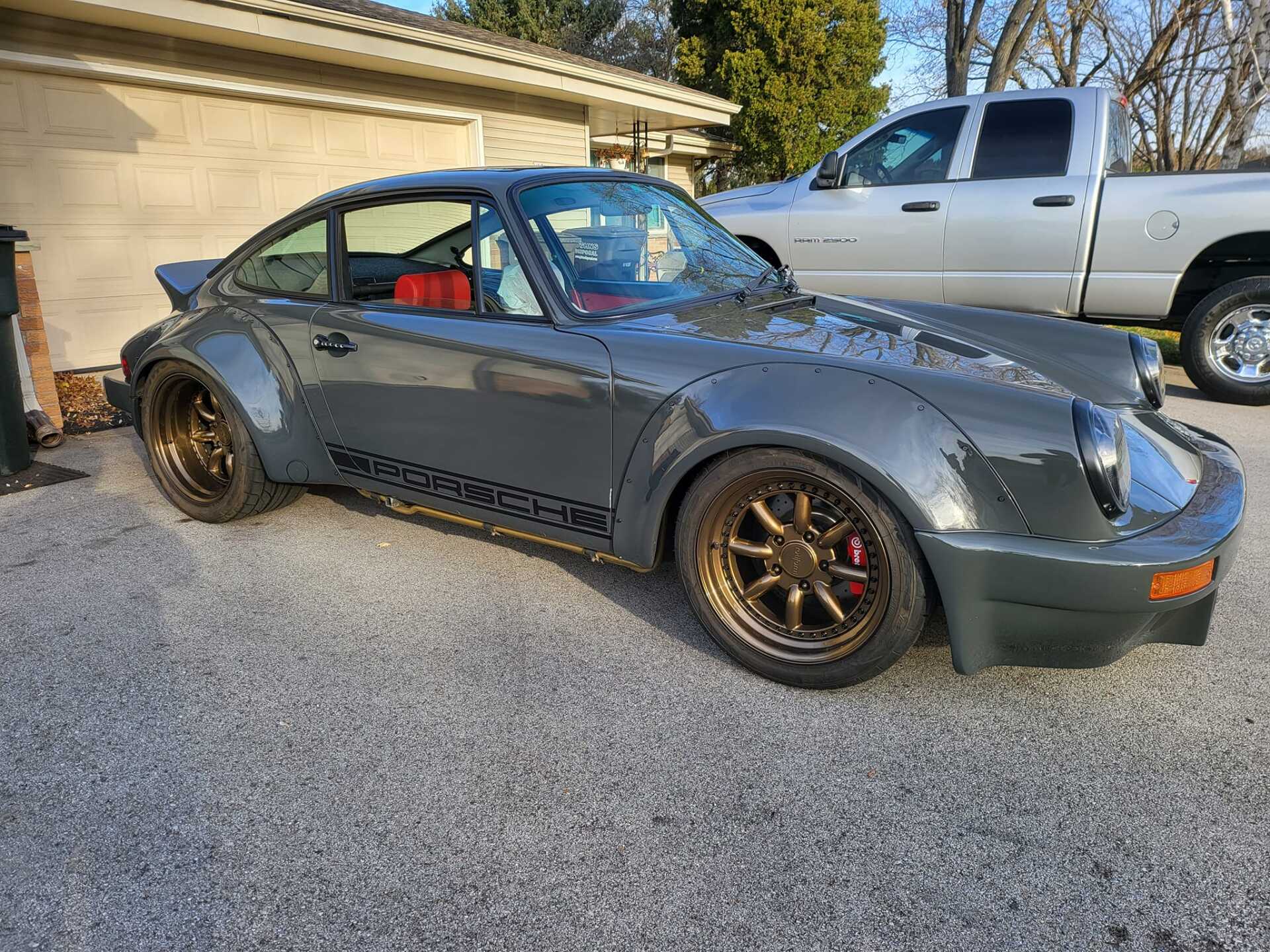 Dark gray Porsche 911 with bronze wheels parked outside a garage, with a pickup truck in the background.