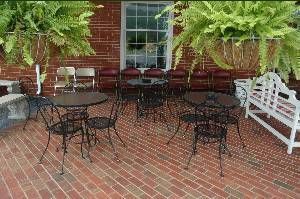 Brick patio with black metal tables and chairs, potted ferns, and a white bench by a brick wall