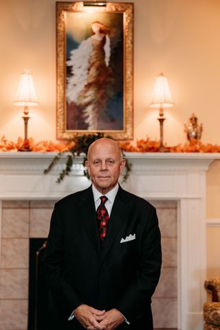 Man in a suit smiles, resting arms on patterned upholstery. Yellow tie, white pocket square. Indoor setting.