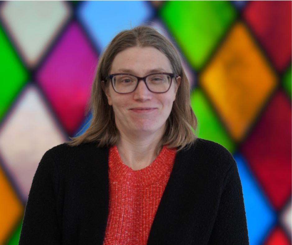 A woman wearing glasses is smiling in front of a stained glass window.