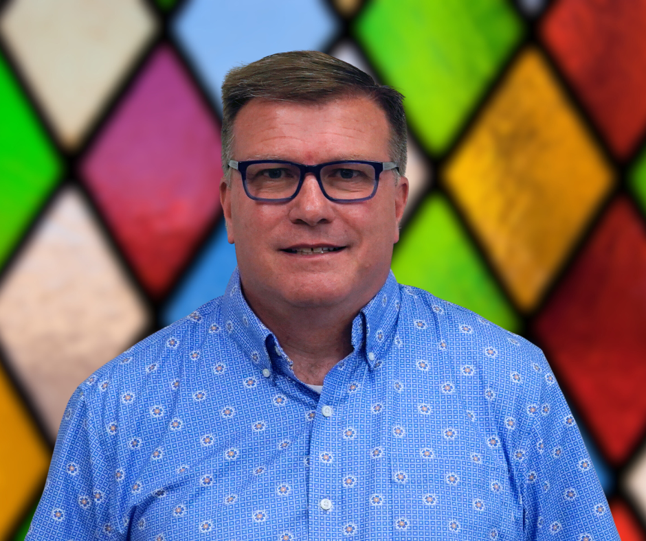 A man wearing glasses and a blue shirt is standing in front of a stained glass window.