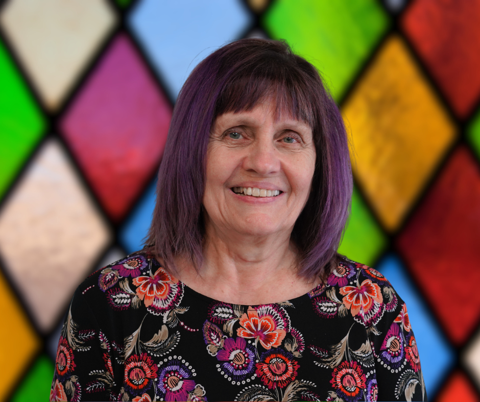 A woman with purple hair is smiling in front of a stained glass window.