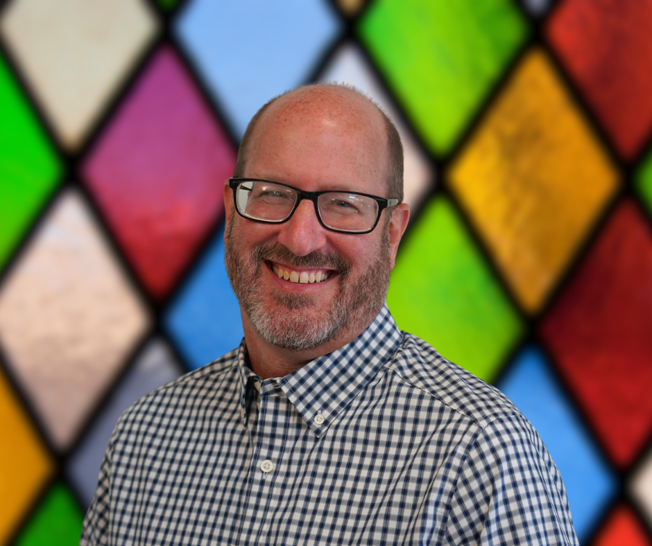 A man wearing glasses and a plaid shirt is smiling in front of a stained glass window.