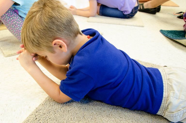 A young boy in a blue shirt is laying on the floor praying.