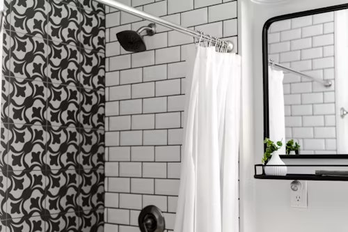 A bathroom with white tiles , a shower curtain , a mirror and a vase.
