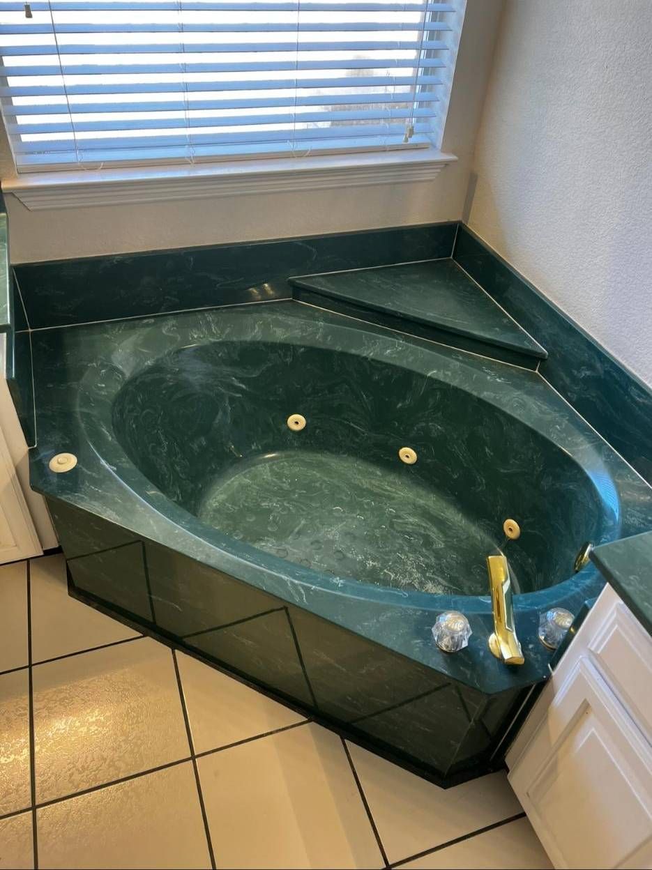 A green marble corner bathtub with gold fixtures and jets, set against a window with white blinds and tiled floors.