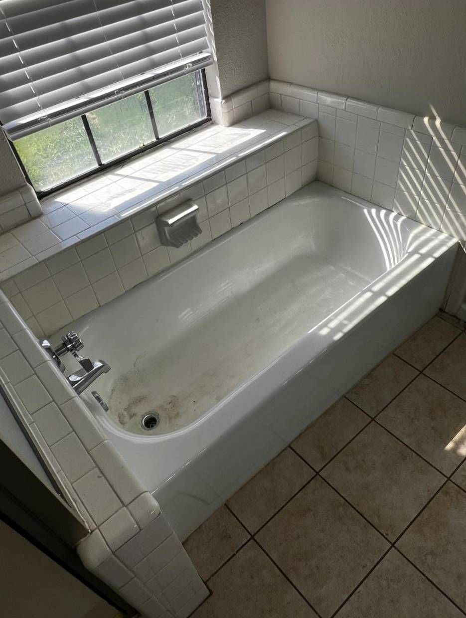 A white built-in bathtub with a tile surround and chrome faucet positioned beneath a window with closed blinds.