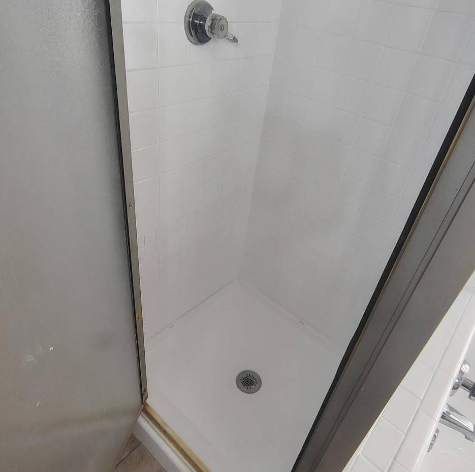 A small shower stall with white tiled walls and a white floor, featuring a central chrome shower handle and drain.