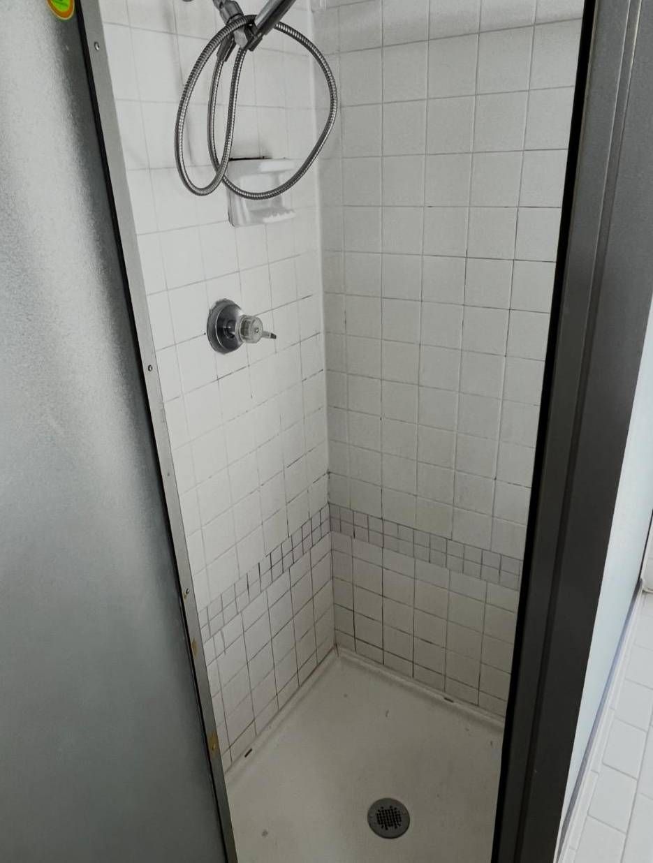 A white tiled shower stall featuring a shower head, handle, and floor drain, framed by a dark doorway.