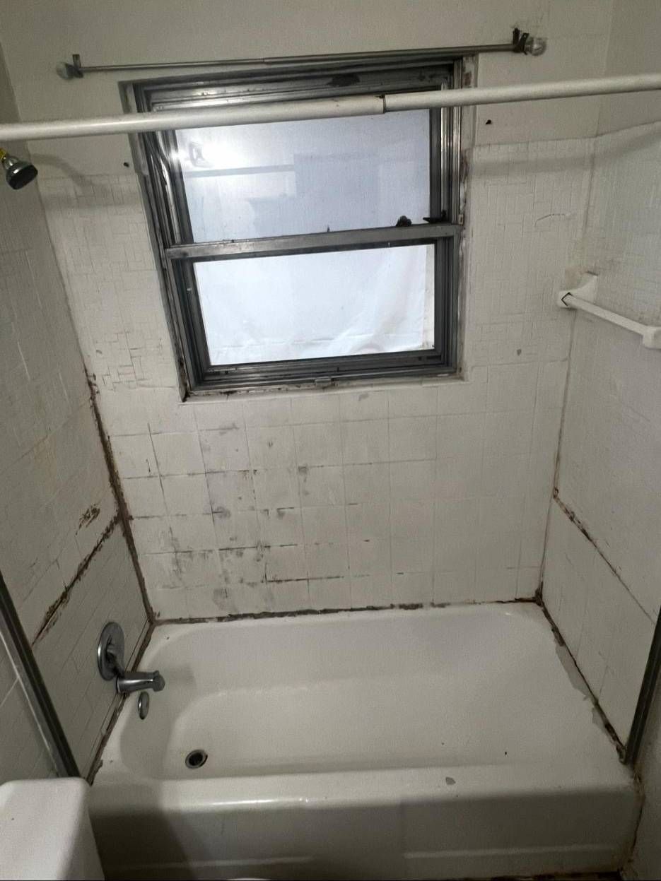A worn bathroom featuring a white bathtub, a window, and tiled walls with visible grout staining and degradation.