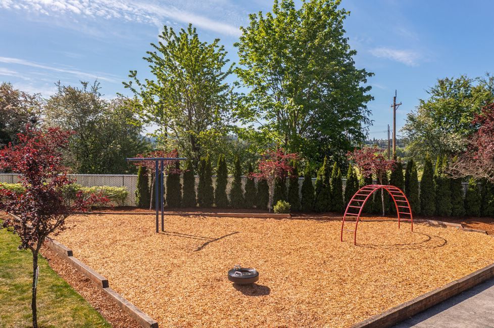 Landscaped playground with tan mulch, trees, shrubs, and a red climbing arch under a blue sky