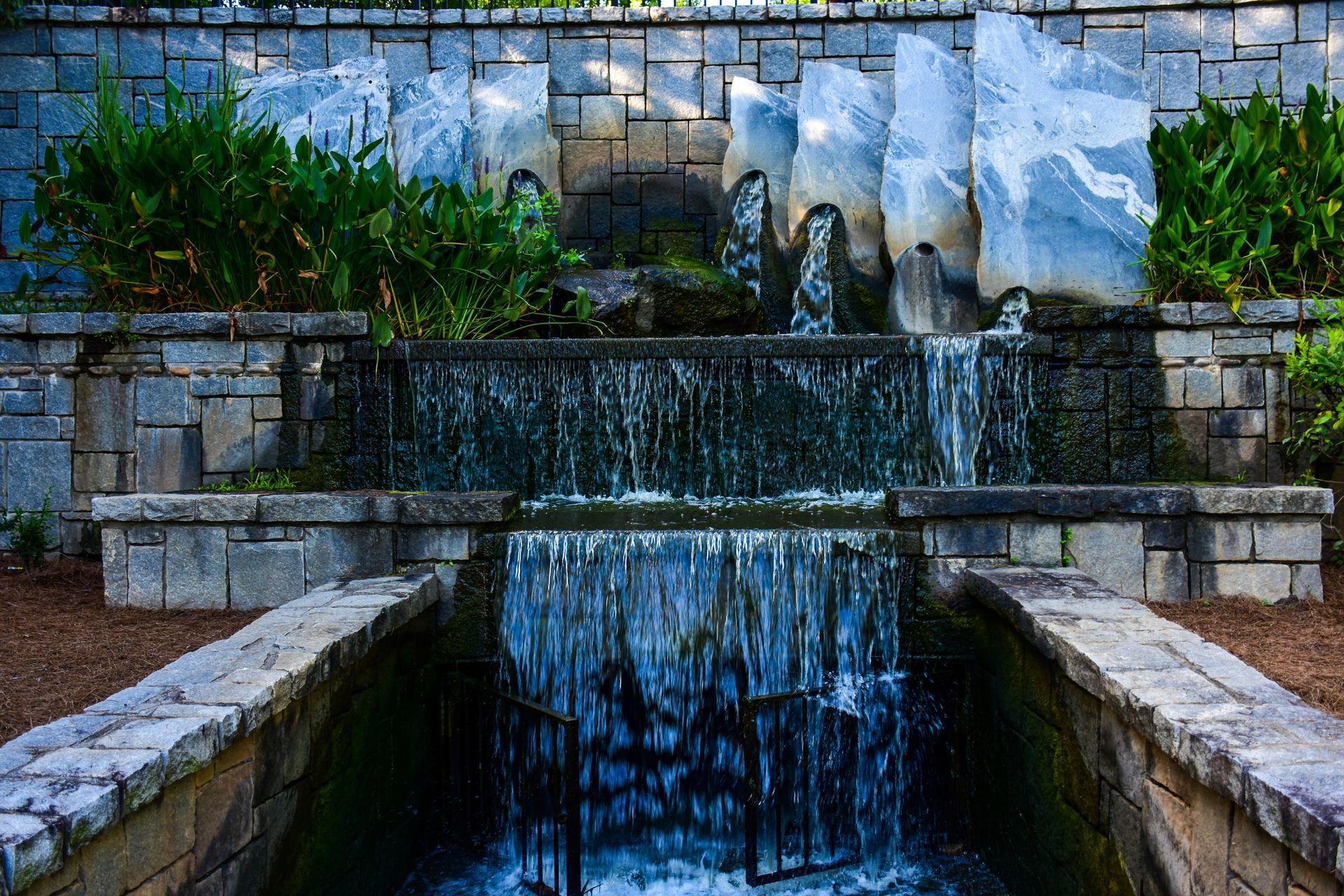 Stone waterfall feature with cascading water into a rectangular pool, flanked by stone walls and greenery.