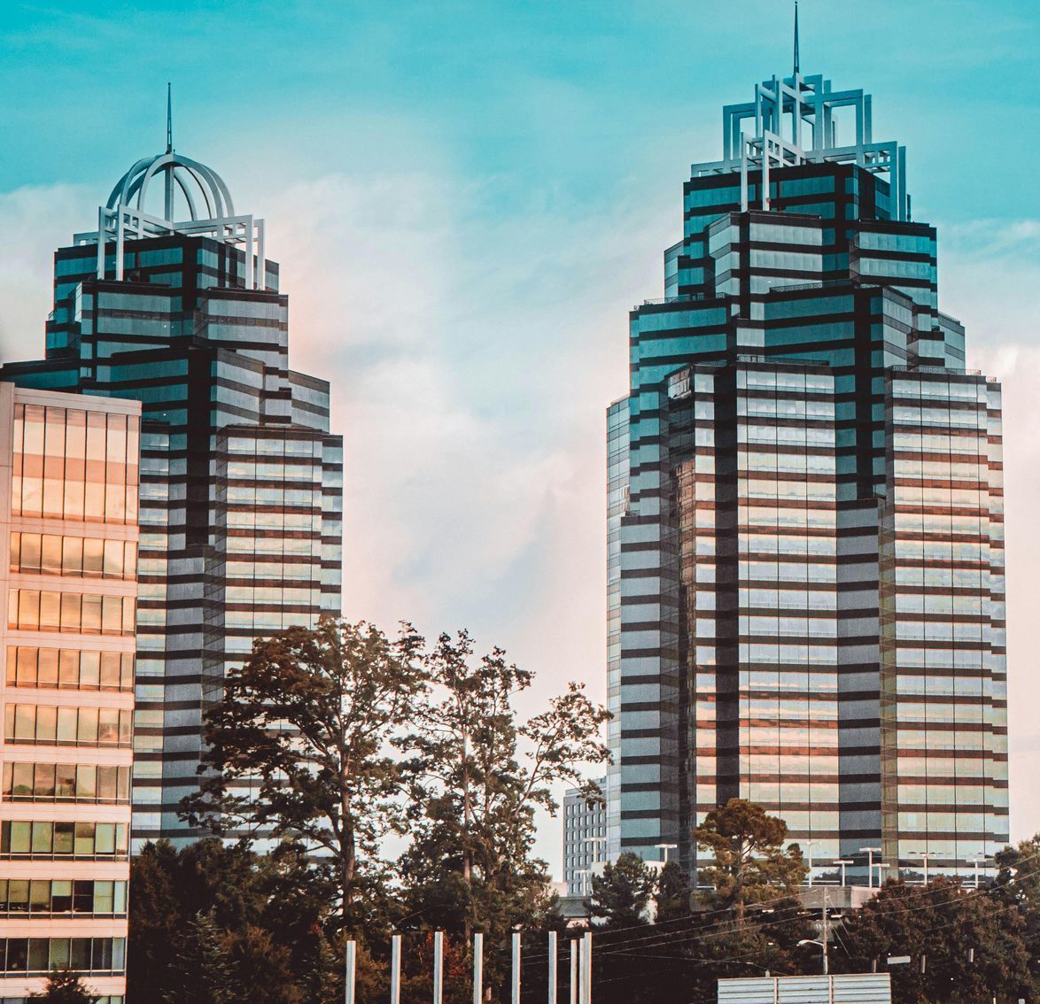 Two tall, modern office buildings with teal glass exteriors and ornate tops, set against a partly cloudy sky.