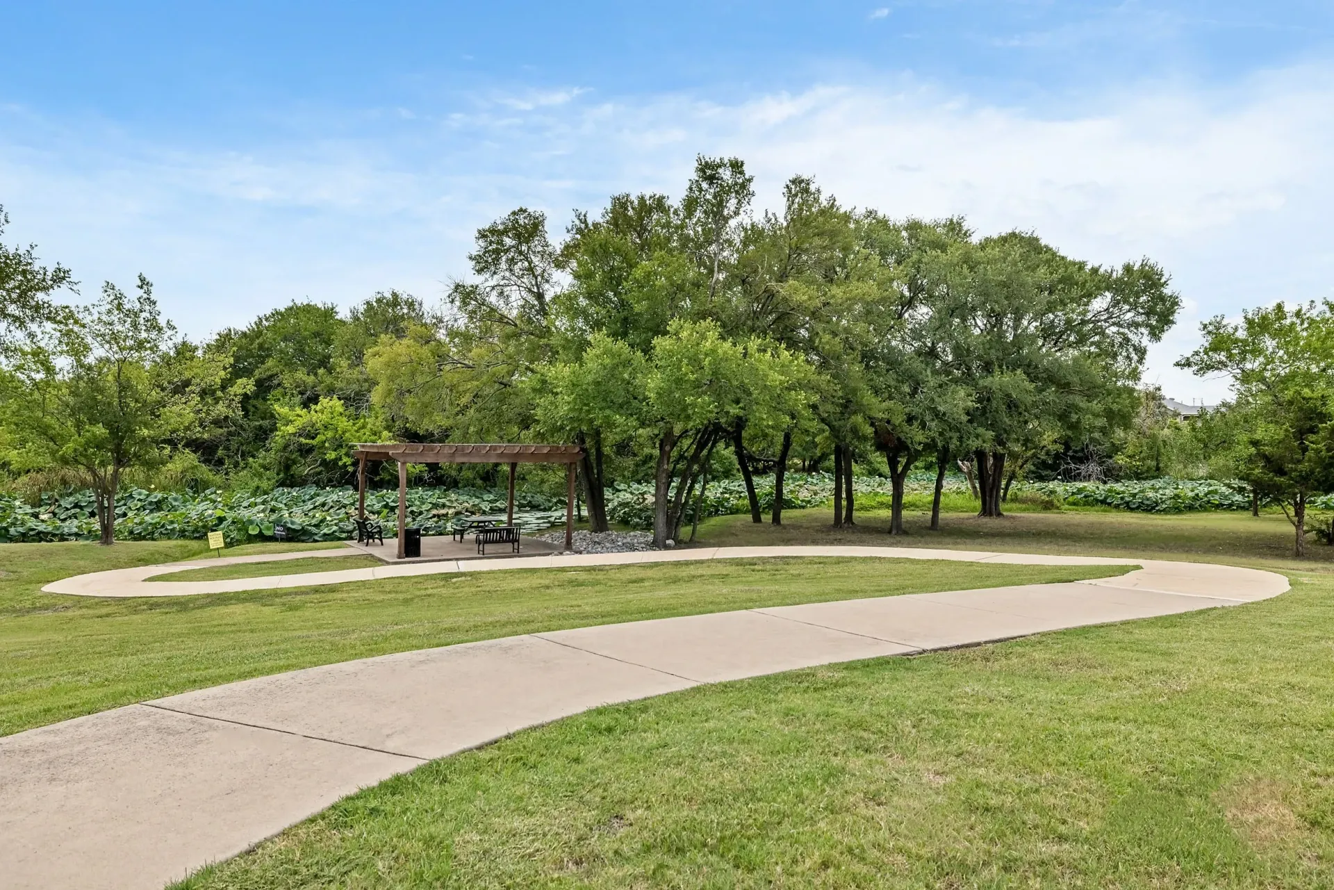 Walking path curves through a grassy area with a picnic shelter and lush greenery in the background.