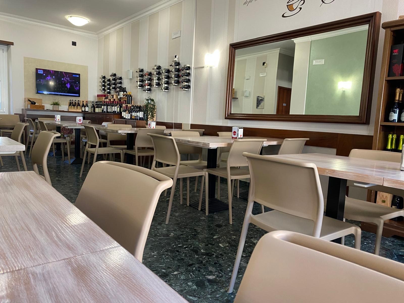 Vista interna di un bar con tavoli e sedie, un grande specchio e bottiglie di vino esposte.