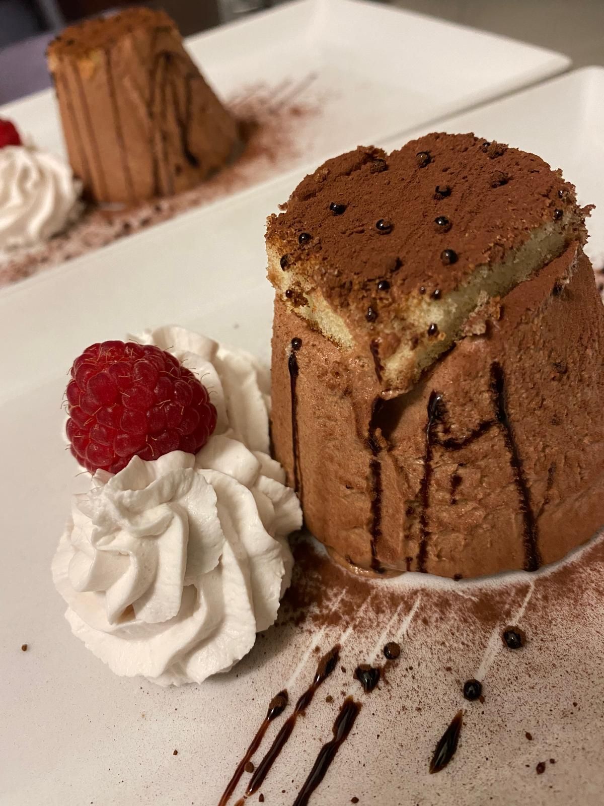 Dessert di mousse al cioccolato con guarnizione di torta, panna montata, lamponi e gocce di cioccolato su un piatto bianco.