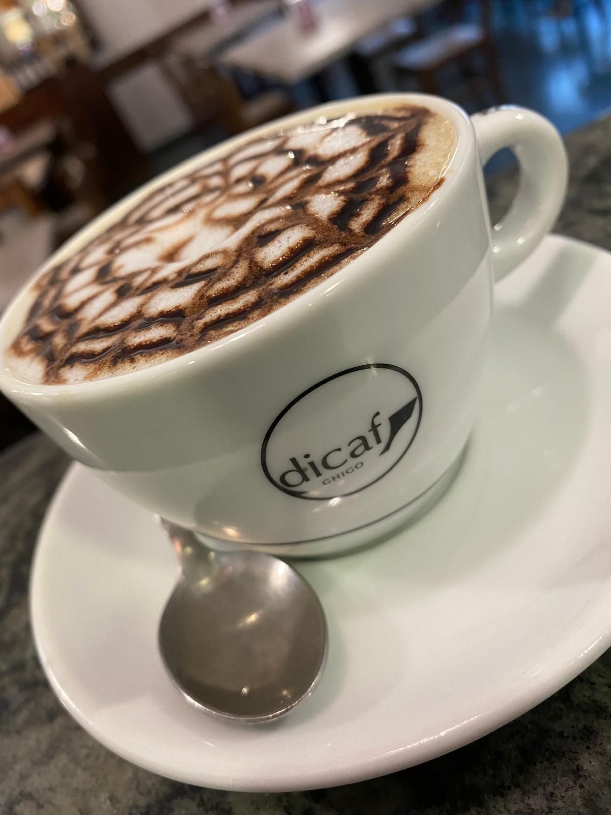 Cappuccino in una tazza bianca, su un piattino, con motivo a spirale di cioccolato; il cucchiaino è appoggiato sul piattino.