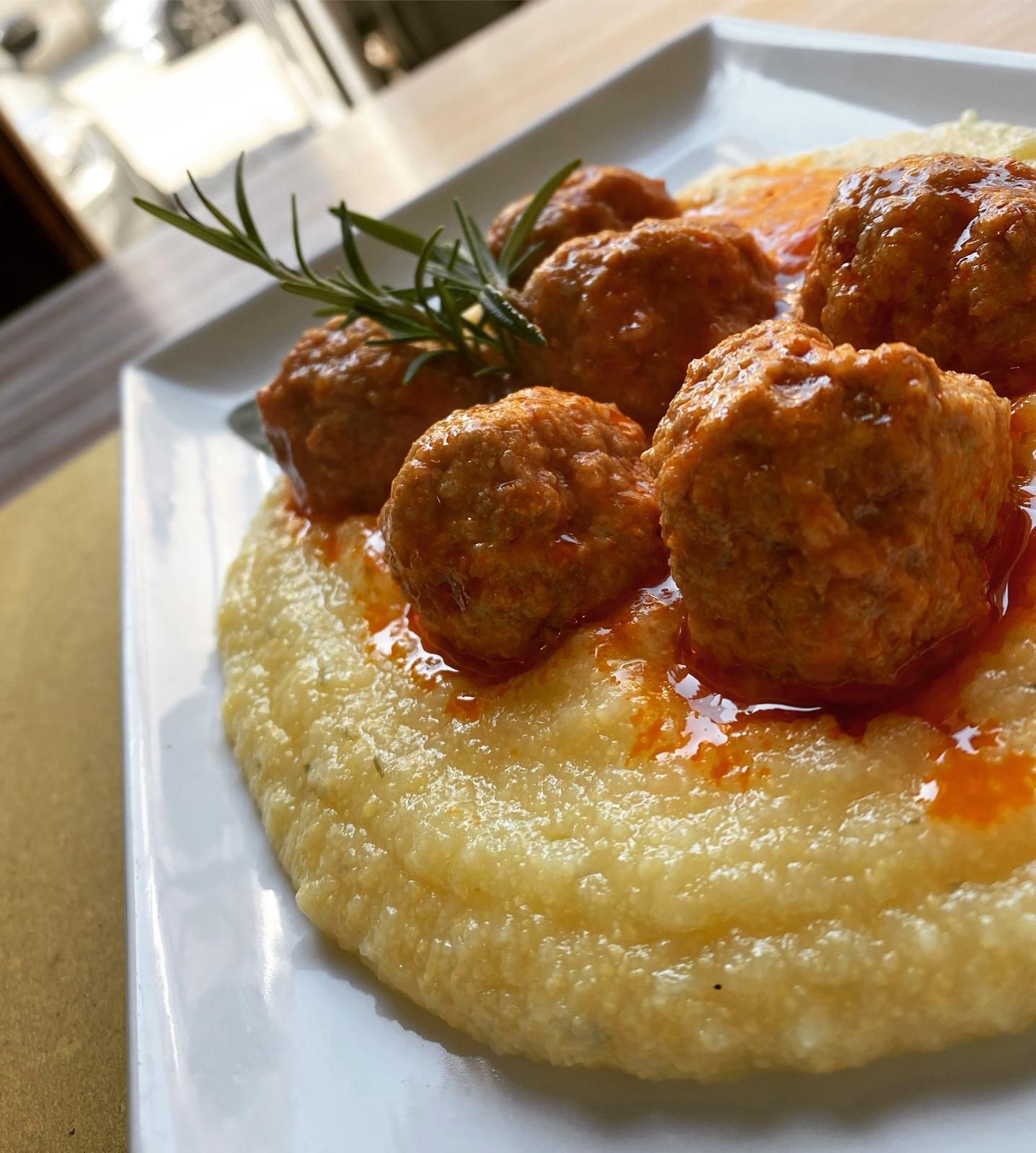 Polpette in salsa di pomodoro servite su polenta, guarnite con rosmarino su un piatto bianco.
