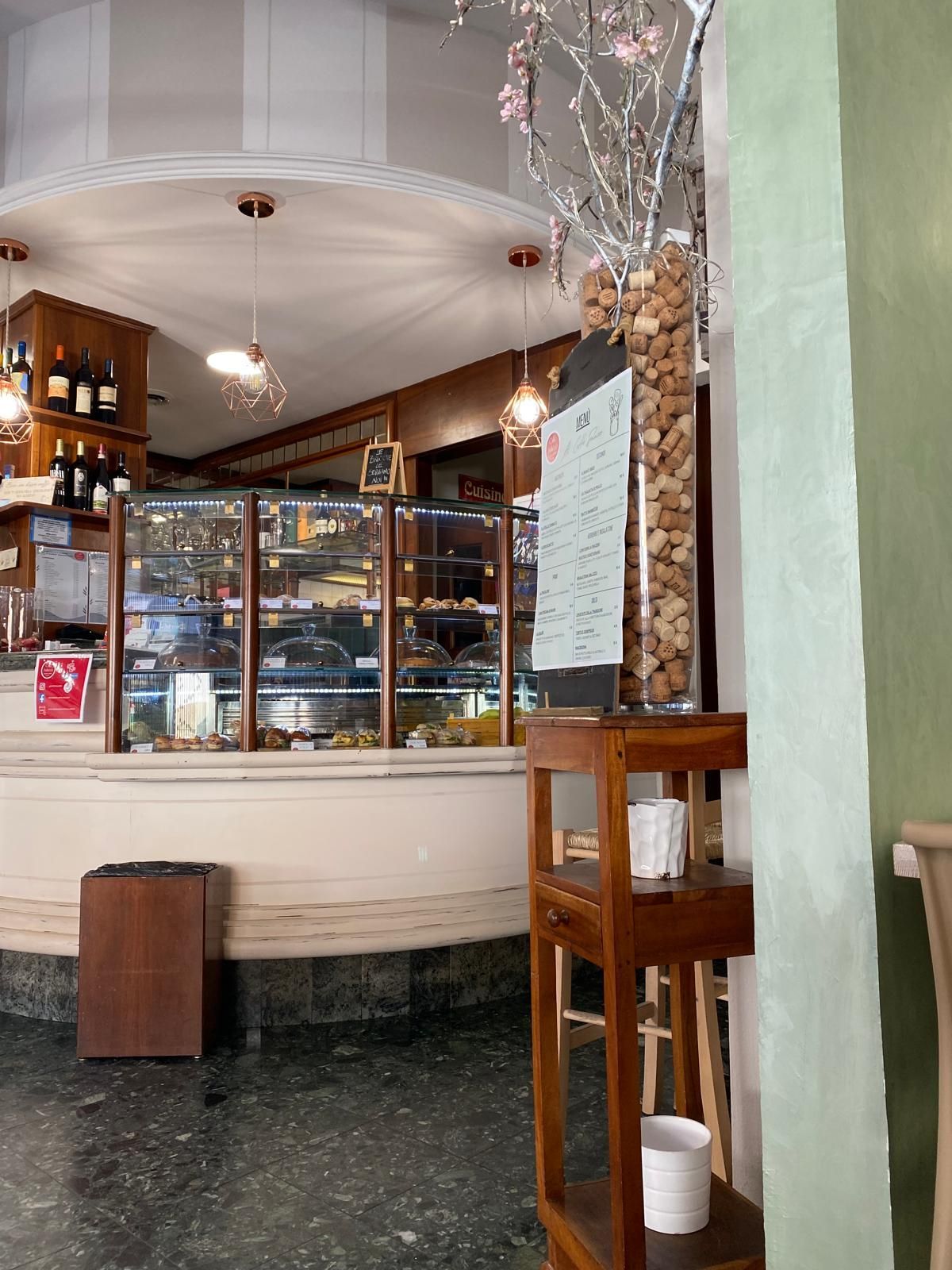 Vista interna di un bar con una vetrina di dolci. Un menù è esposto su un tavolo di legno.