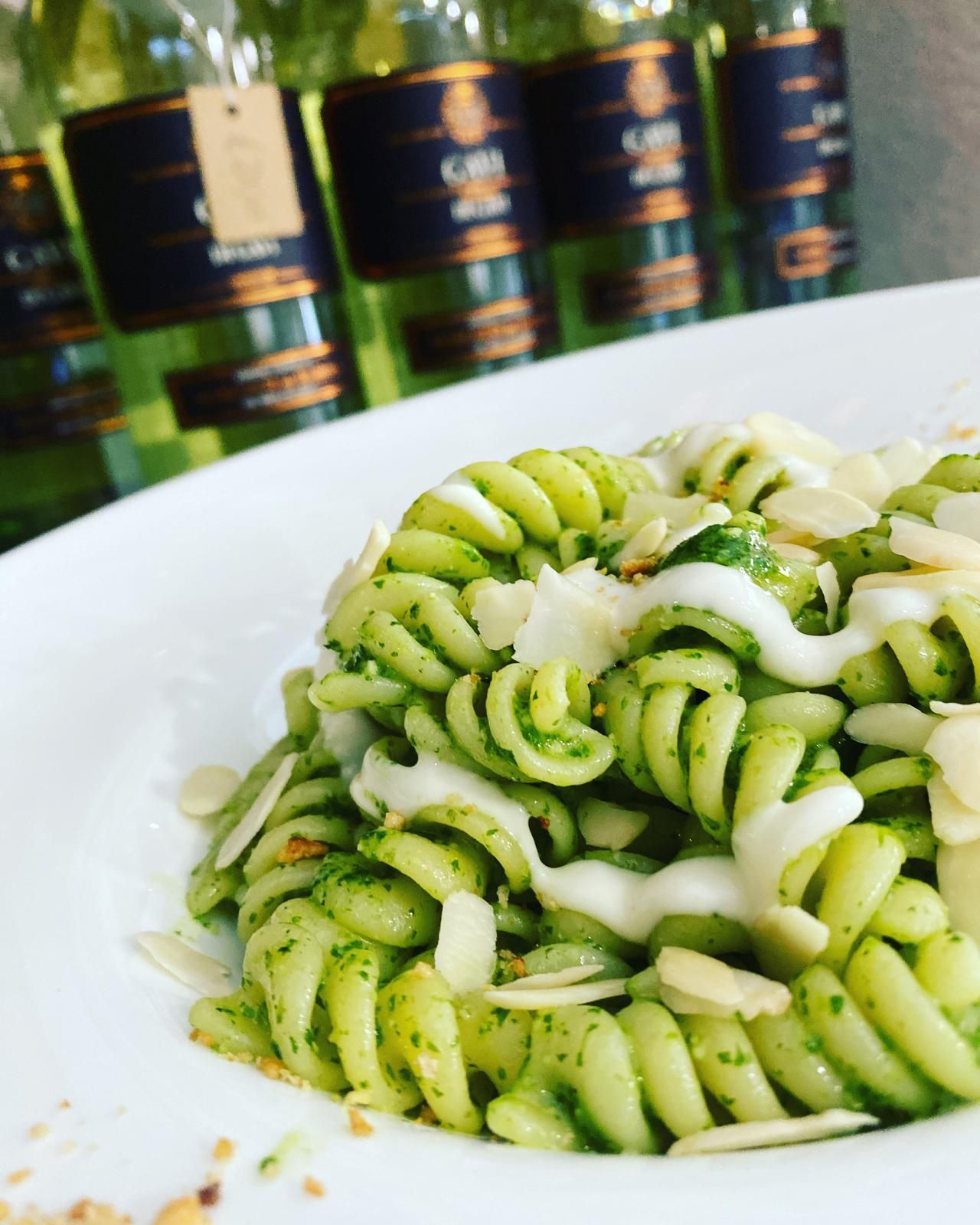 Pasta al pesto verde con panna e mandorle, piatto bianco, bottiglie di vino sullo sfondo.
